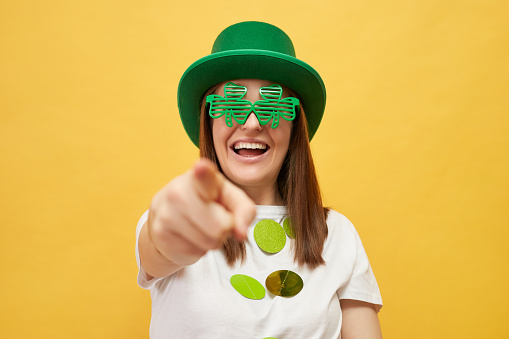 National holiday fun. St. Patrick's Day greetings. Smiling woman wearing festive green hat and shamrock glasses standing isolated over yellow background pointing at camera