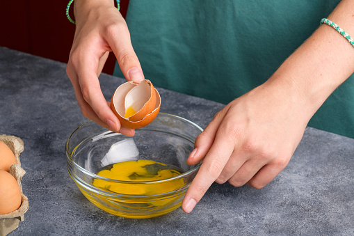 A woman's hand holding the eggshells of breked egg into a clear glass bowl in the gray kitchen table, the process of cooking, fresh eggs into the bowl is ready to cooking, Lifestyle, close up
