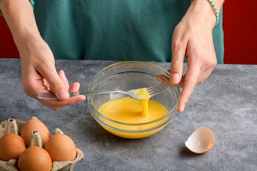 Female hands beat eggs with fork in a glass bowl on a gray kitchen table. Cooking scrambled fresh eggs for breakfast, the process of omelette cooking, close up