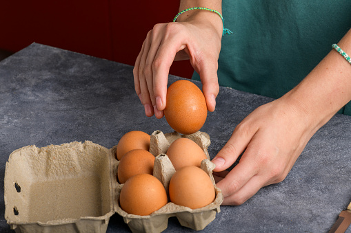 A woman's hand holding an egg taked from the egg box that is on the gray kitchen table, close up, food lifestyle, ready to cooking