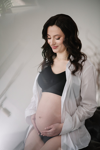 A pregnant woman holds her belly sitting on the sofa at home. Young woman in a light room. Pregnancy motherhood concept. Female waiting for baby. Pregnant girl touching belly, caring health indoors.