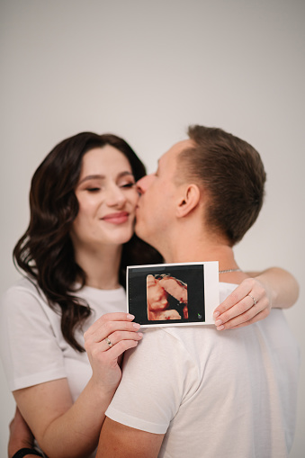 Pregnant couple holding in hands an ultrasound scan of their baby. Happy family pregnancy, expectation. Male hugs female closeup. A man, husband hugging his pregnant wife in the home.
