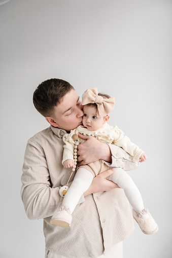Portrait of daddy kisses an adorable baby girl with bow in hair isolated in white background. Cute baby in dad's arms closeup. Dad holds in hands a 6 month old little girl. Father hugging his daughter