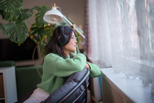 Part of a series of daily life of a woman at home, using headphones next to window.