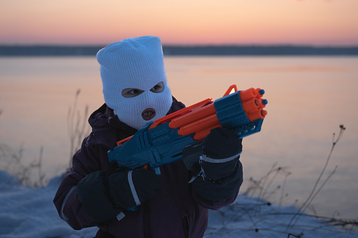 A child, a little girl in a white balaclava, plays a war game with tiny plastic weapons at sunset in winter. High quality photo