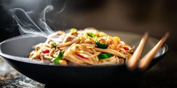 Close-up shallow depth of field photo of stir-fried noodles with vegetables and tofu on a black plate with chopsticks.
