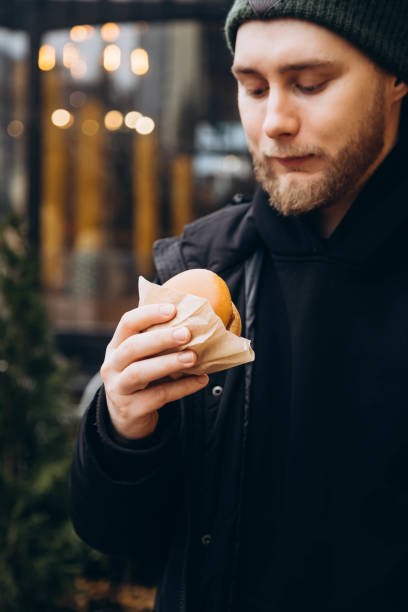 istock Attractive bearded young man wearing a jacket and hat with a burger outside. 2078389857