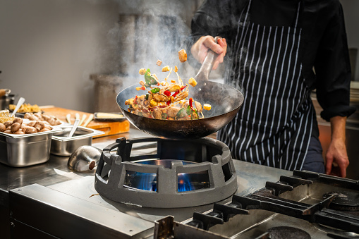 Unrecognizable male chef tossing vegetables and meat in the air while stir frying in a wok in a professional kitchen.
