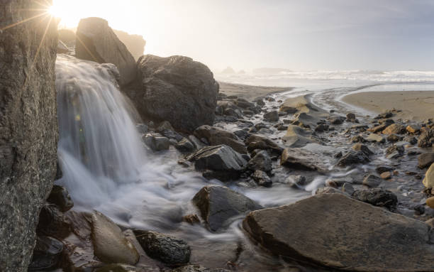 A stream of water flows between large rocks on a beach. The water is clear and calm, and the rocks are scattered throughout the scene