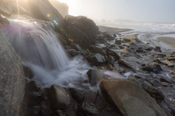 A waterfall is flowing into a river on a rocky beach. The water is clear and the rocks are large. The scene is peaceful and serene