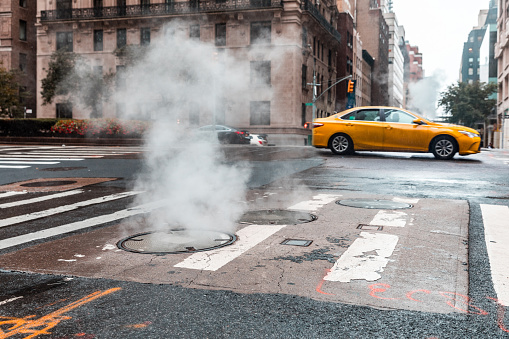 Vibrant urban street with steam rising from a manhole and a taxi in motion in New York - Steamy street scene with yellow taxi in the city