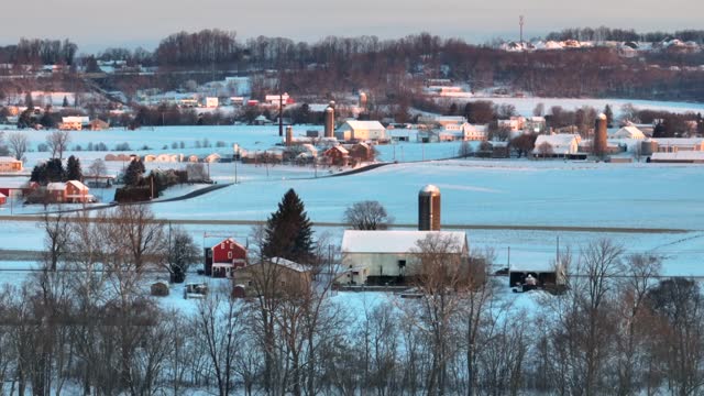 Farmland in rural USA covered in snow during winter. Aerial view of farms and fields with silos, barns, and houses.