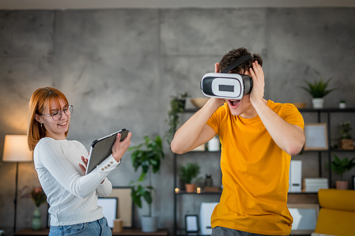Two young students having fun with virtual reality headset at home, one woman holding digital tablet while a man wearing virtual reality headsets