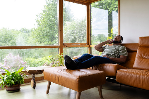 A mid adult man listening to music through wireless headphones and sitting with his feet up in his living room at home in Hexham, North East England. He is holding onto the headphones and looking content with his eyes closed.