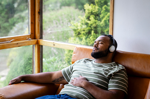 A mid adult man listening to music through wireless headphones and sitting back on a leather sofa in his living room at home in Hexham, North East England. He is listening to the music while looking content with his eyes closed.