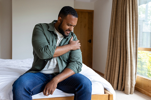 A mid adult man sitting on the edge of his bed in the bedroom at home on a morning in Hexham, North East England. He is looking in distress as he feels his shoulder, which is causing him pain due to injury.