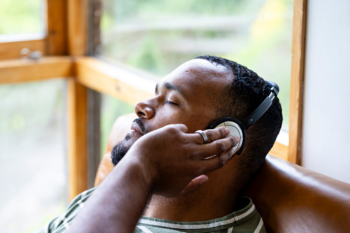 A mid adult man listening to music through wireless headphones and sitting back on a leather sofa in his living room at home in Hexham, North East England. He is holding onto the headphones and looking content with his eyes closed.