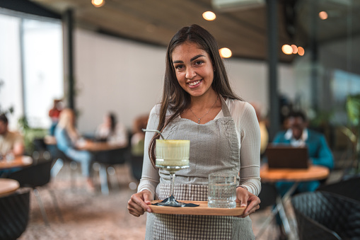 Portrait of a young adult Hispanic waitress in a modern cafeteria, exuding excellence as she presents a delightful matcha latte on a stylish serving tray. Her attention to detail and commitment to providing an exceptional experience are evident in the way she carefully holds the tray, ensuring that every aspect of the presentation is flawless. This tasteful display of professionalism enhances the enjoyment of the matcha latte, making it a truly memorable and satisfying moment for the customers in the modern cafeteria.