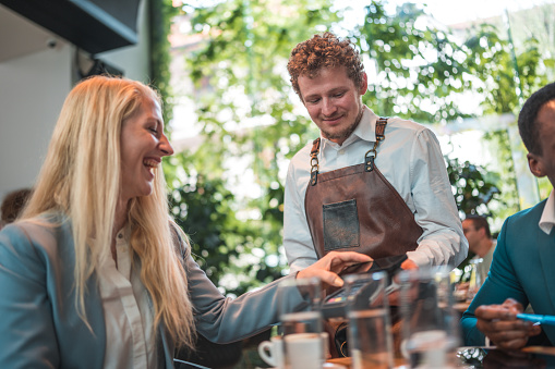 Witness the seamless integration of technology in business interactions as diverse partners meet in a modern artisan coffee house. Leading the way in contactless payments, a tech-savvy Caucasian female expertly uses her smartphone, exemplifying the efficiency and convenience that technology offers in a diverse business setting.