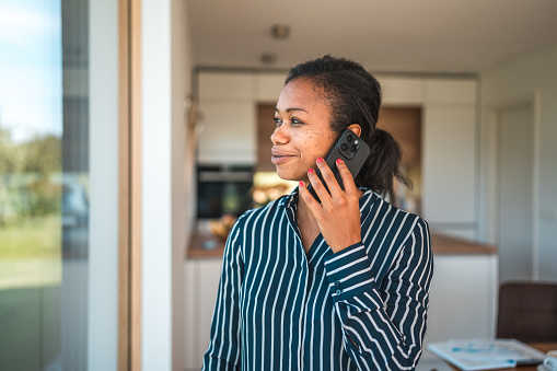 Waist up image of a self-employed multiracial casting agent in striped shirt, looking out the window  while chatting  on the mobile phone, an open notebook on the dining table behind her and a view of the kitchen.