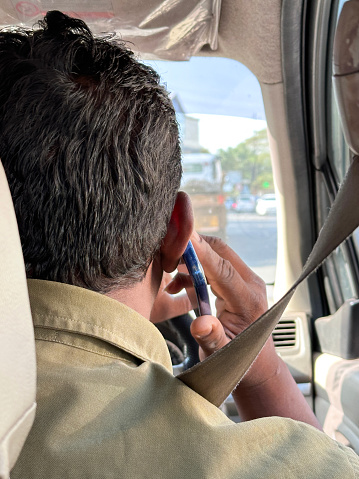 Stock photo showing an unrecognisable man driving on a busy motorway in his car (a private hire taxi cab car), whilst talking to a friend on his mobile phone. The highway is visible through the windscreen and windows, while the driver is pictured with the smart phone being held to his ear as his listens and drives, wearing a seatbelt across his chest. This is a concept image for dangerous driving, since using a phone behind the wheel is illegal and considered to be both dangerous and a serious traffic offence, punishable with large fines and points on the relevant driving licence.