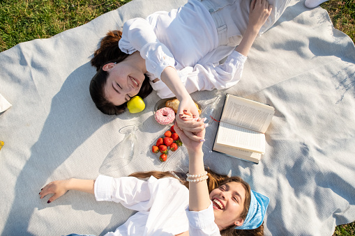 Young women relax and shake hands lying on white blanket on grass. Best girlfriends laugh and relish summer picnic together