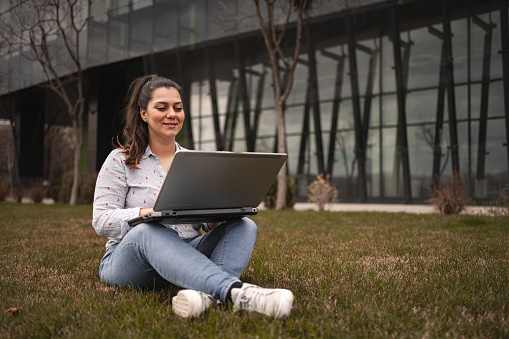 Woman outdoors sitting on grass working on laptop.