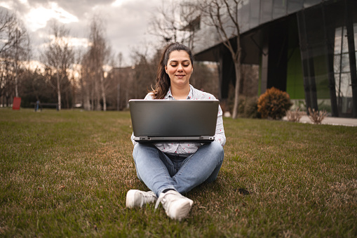 Woman outdoors sitting on grass working on laptop.