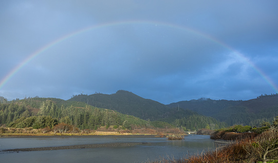 A rainbow is seen over a body of water. The sky is cloudy and the water is calm