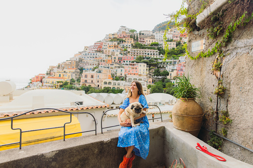 Smiling female with long hair sitting with her pug with scenic view of old cliffside buildings of Positano city in Amalfi Coast