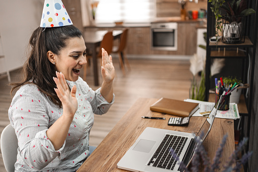 Excited woman with party hat talking on a video call over laptop at home.