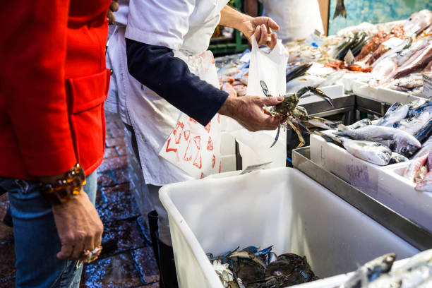 elegant lady at market to fish counter - the seller is putting callinectes sapidus blue crabs into the bag - blue crabs are american atlantic coasts - they cause damage to fishing by preying on clams - prepared shellfish tray variation catch of fish imagens e fotografias de stock