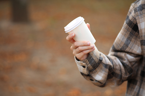 Woman in warm shirt holds White cardboard cup for hot drink (coffee, cup) outdoors in autumn park