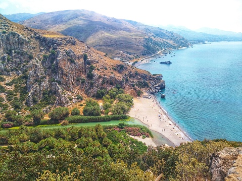 A captivating view from the cliff capturing a mountain river winding through a palm grove and flowing into the sea. The shore reveals a beach amidst rocks, offering a stunning panorama in Crete
