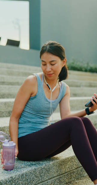 istock Young Asian female athlete runner drinking protein shake and using smartphone after exercise workout in urban park. Diet and healthy food workout exercise. Vertical Screen. 2078797297