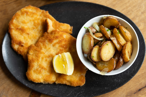 Wiener schnitzel with fried potato and sweet potato in black plate on a wooden table.