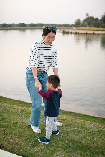 A mother and her young child share a sweet moment at their lakeside picnic, dancing and having fun surrounded by the serene beauty of nature. Location: Love lakes near Al Qudra in Dubai, United Arab Emirates