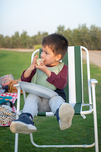 A young boy sits comfortably in a green and white striped picnic chair, enjoying a sandwich outside with a natural backdrop of a lake on a camping trip. Location: Love lakes near Al Qudra in Dubai, United Arab Emirates