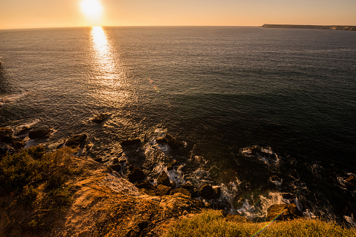 Sunset at cliffs and Atlantic ocean hitting rocks in Sagres, Algarve, Portugal. Lighthouse at the Cabo de São Vicente in the distance.