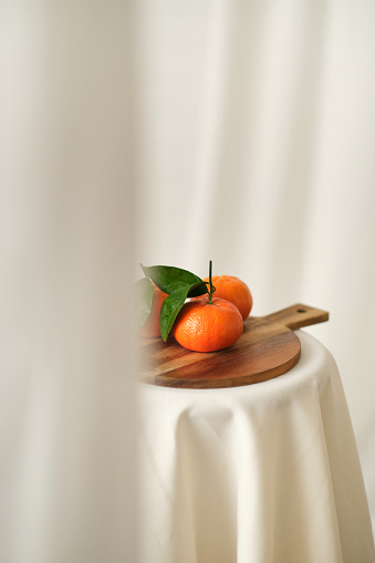 Vertical still life studio photo of a white curtain partially open with oranges in a wooden board
