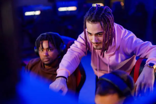 Portrait of a young cybersport gamer with dreadlocks pointing at the screen while discussing strategy with his diverse team in cyber club while playing a video game. Esport streaming game online.