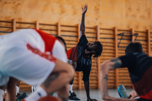An african american professional basketball player is stretching his backs on court with his teammates during the training. Interracial basketball player is warming up for training on basketball court