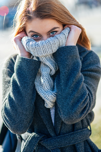 Portrait of Happy young woman holding woolen scarf with hands over nose to protect from the frost. Portrait of beautiful young woman in warm winter knitted clothes covering her face and looking at camera.