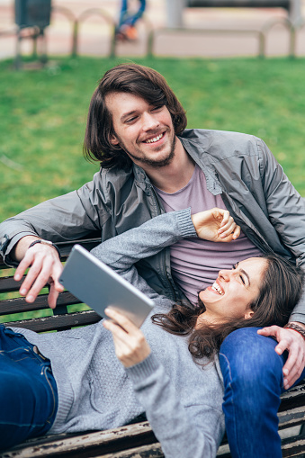 Happy young couple using digital tablet outdoor in the city park. Cheerful man and woman on date sitting on the bench taking selfie, surfing the net or text messaging