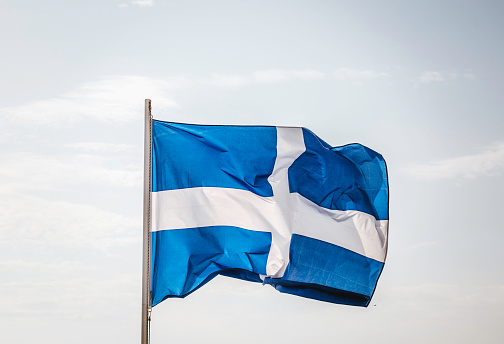 An image capturing the Greek flag fluttering in the wind against a clear blue sky.
