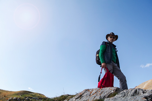 Mature man with red backpack is standing on the mountain peak.  Adult with a hat on his head looks down