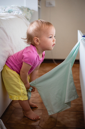 side view of little baby girl with blonde hair pooling t-shirt from the drawers, playing with clothes in the chest