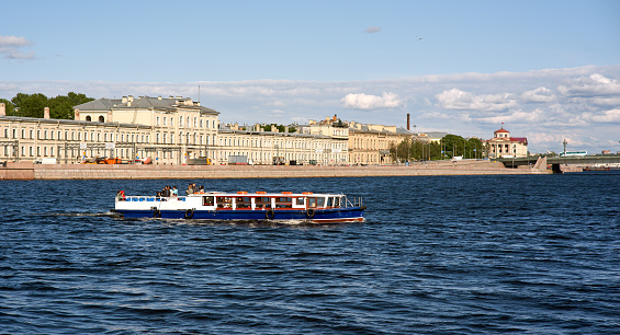 Saint Petersburg. Russia - June 02, 2023: Cityscape of old history buildings on embankment Neva river and tourist boat in the foreground. Blue cloudy sky.