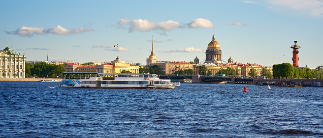 Saint Petersburg. Russia - June 02, 2023: Cityscape of old history buildings on embankment Neva river, Saint Isaac's Cathedral and Rostral column (lighthouse) on Spit of Vasilievsky Island, Saint-Petersburg, Russia. Neva River and tourist boat in the foreground. Blue cloudy sky.