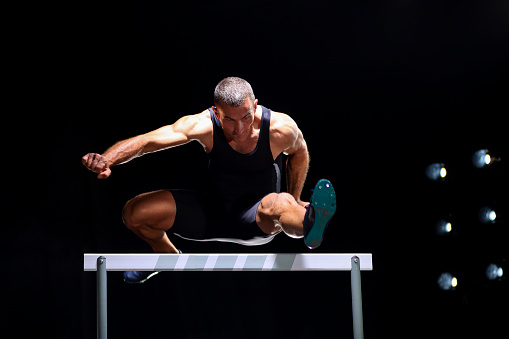 Mid-air shoot of a young man jumping over hurdle in a track race. Stadium light in the back.